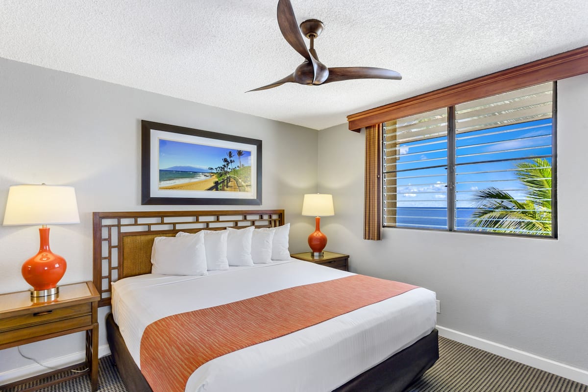 A hotel room with a bed, two bedside tables, a ceiling fan, a window with blinds, and a framed picture on the wall.