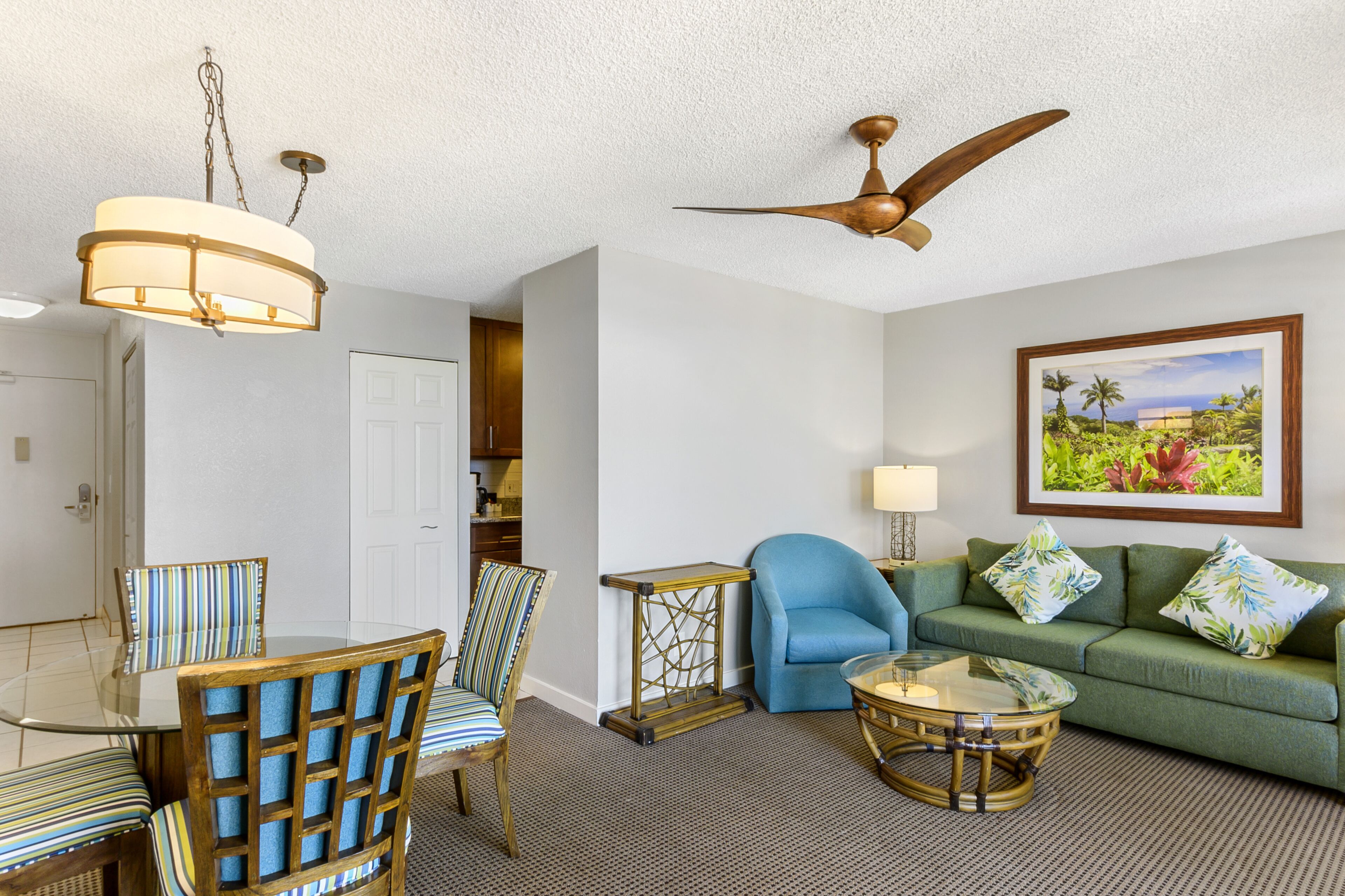 A living room with a glass coffee table, striped chairs, a blue armchair, a green sofa with floral cushions, a ceiling fan, and a framed picture on the wall.