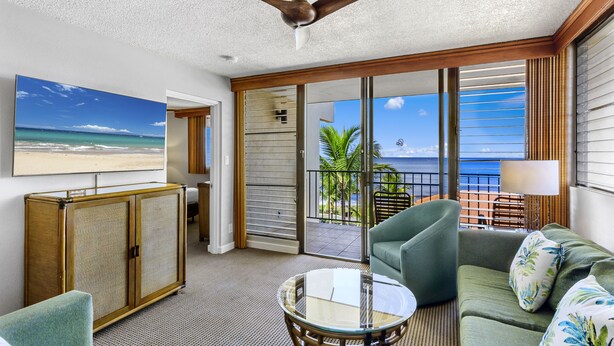 A room with a ceiling fan, a large window with a view of the ocean, a sofa, a table, and a cabinet. - Maui Beach Vacation Club (Kihei)