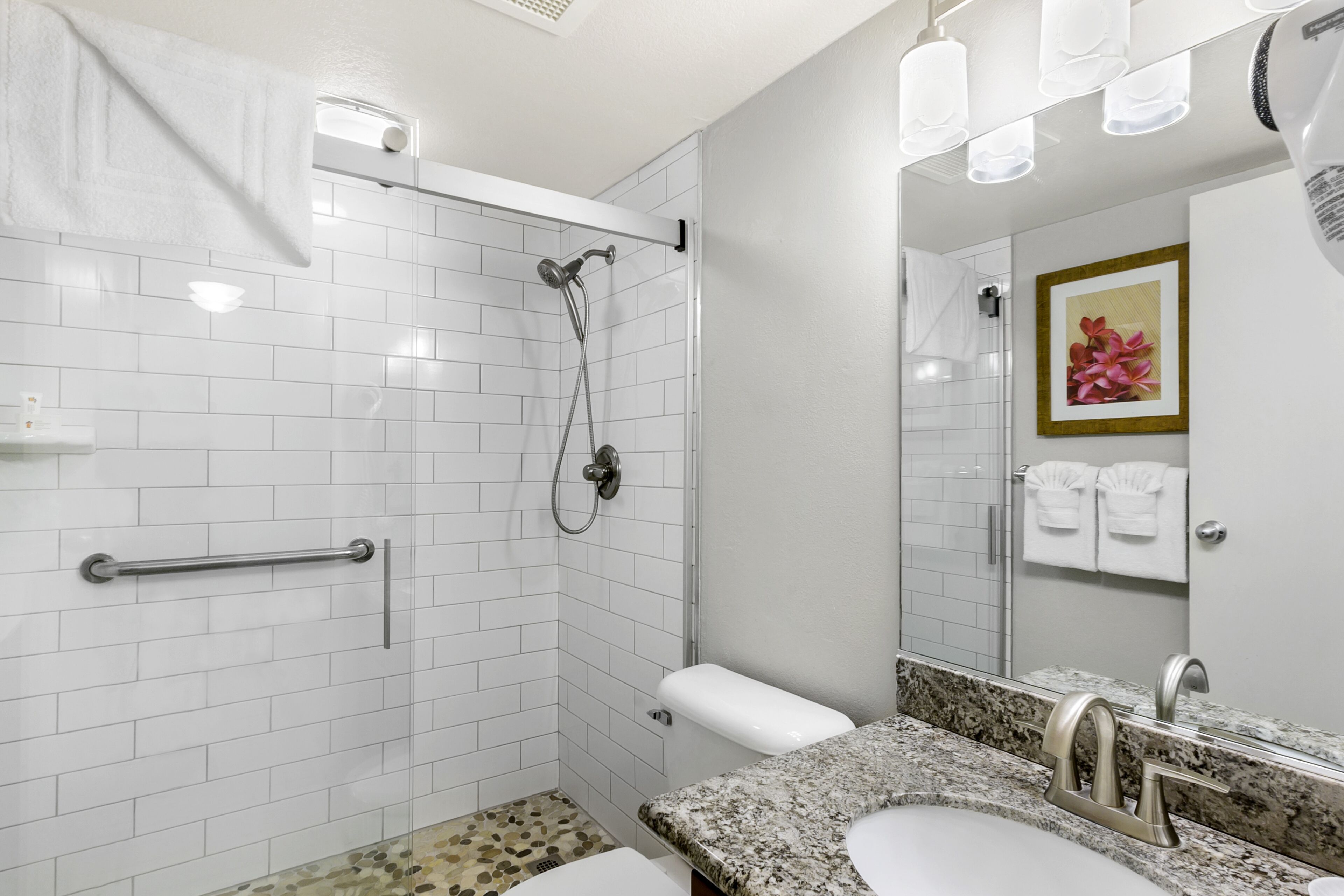Une salle de bain moderne avec des murs recouverts de carreaux blancs, un lavabo sur un plan de travail en granit, une douchette à main et un porte-serviettes.