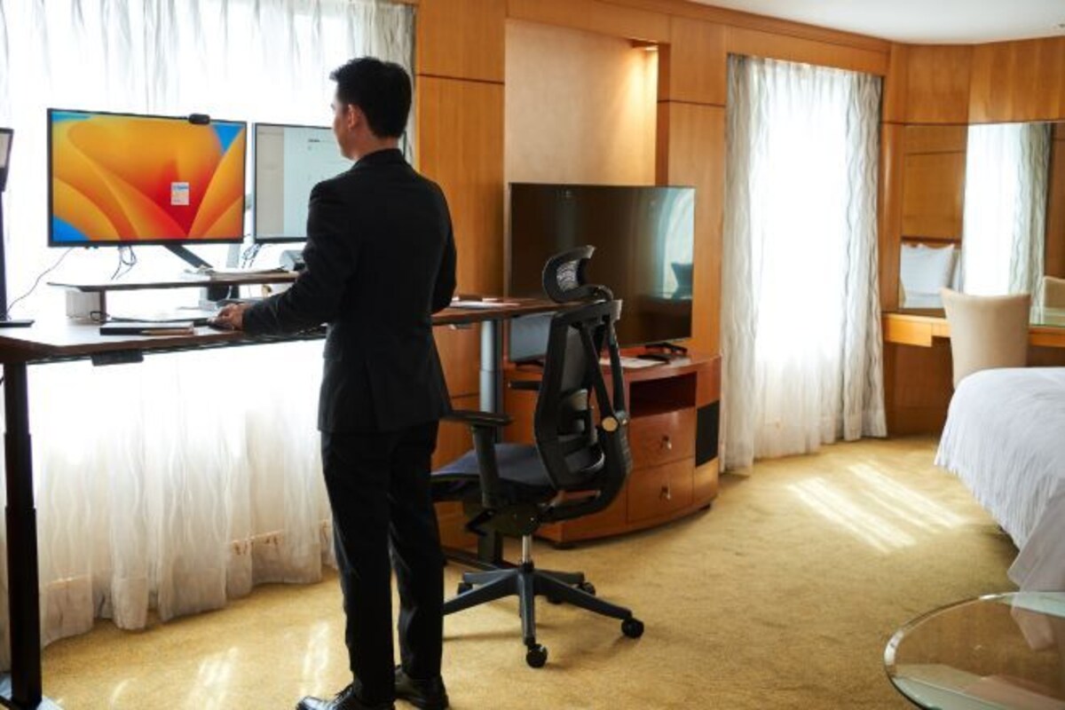 A hotel room with a desk, two monitors, a television, and a bed.