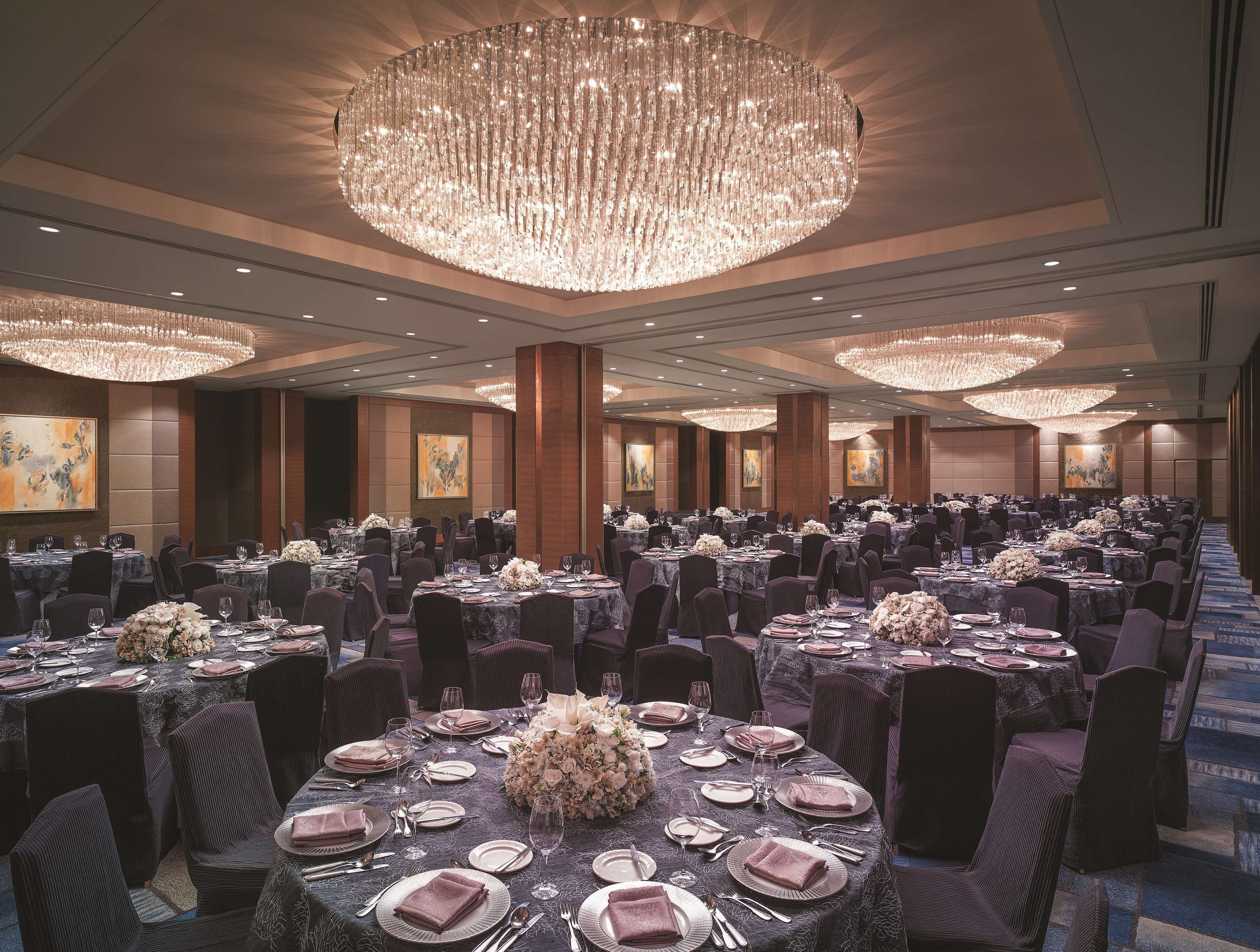 A banquet hall with round tables set for dining, large chandeliers, and artwork on the walls.