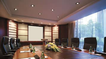 Un salón de conferencias con una gran mesa de madera, varias sillas, una pantalla de proyector y un ventanal con vistas a árboles.
