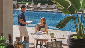 Zona de piscina con mesa y sillas, un camarero sirviendo una bebida y una piscina al fondo.