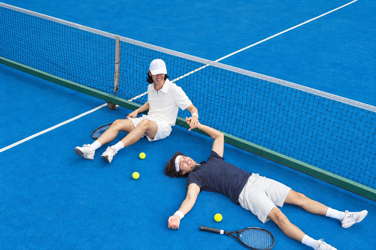 Una cancha de tenis con una red, dos jugadores tirados en el suelo y pelotas de tenis esparcidas.