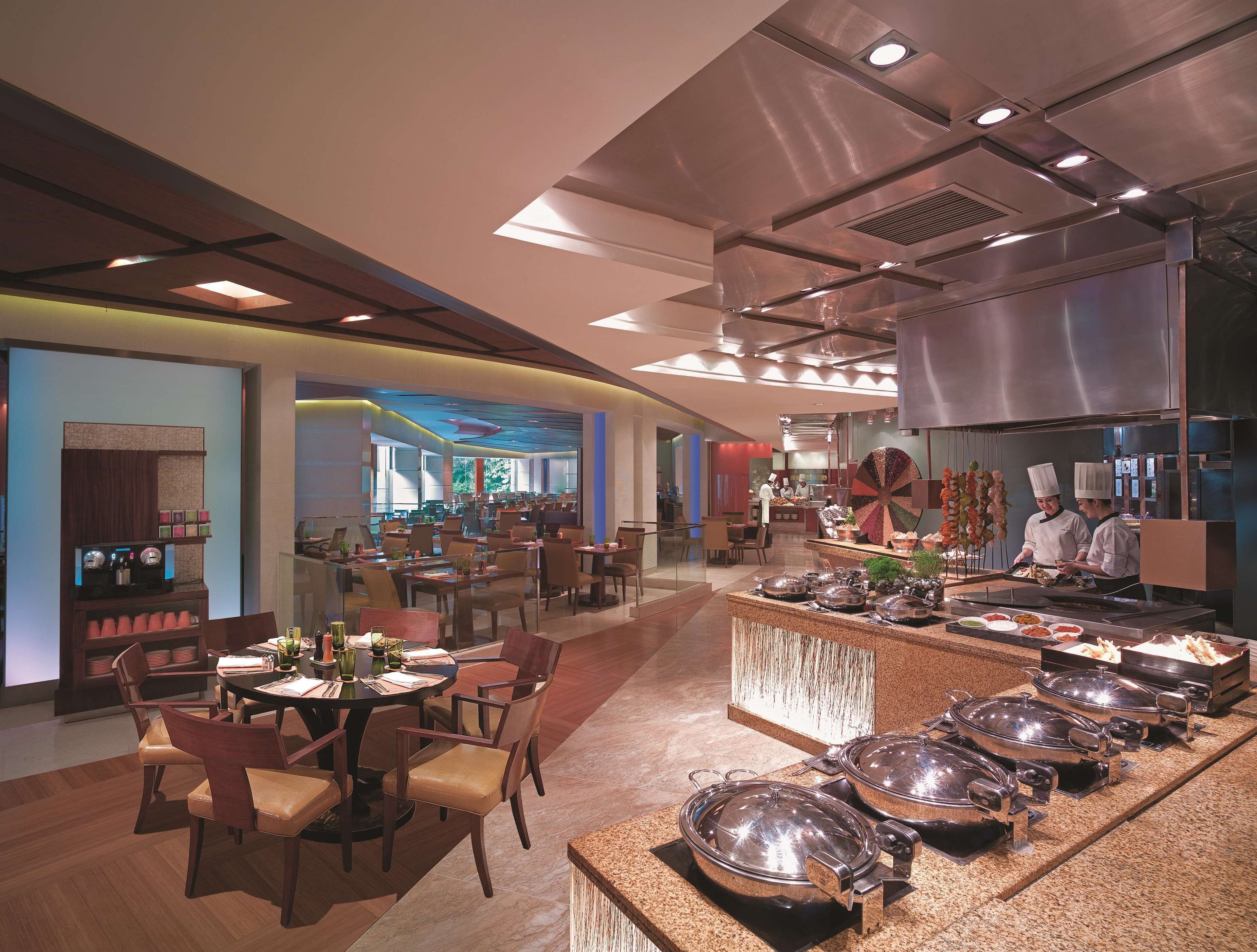 A modern restaurant with a buffet setup, including multiple serving stations and a central dining area with round tables and chairs.