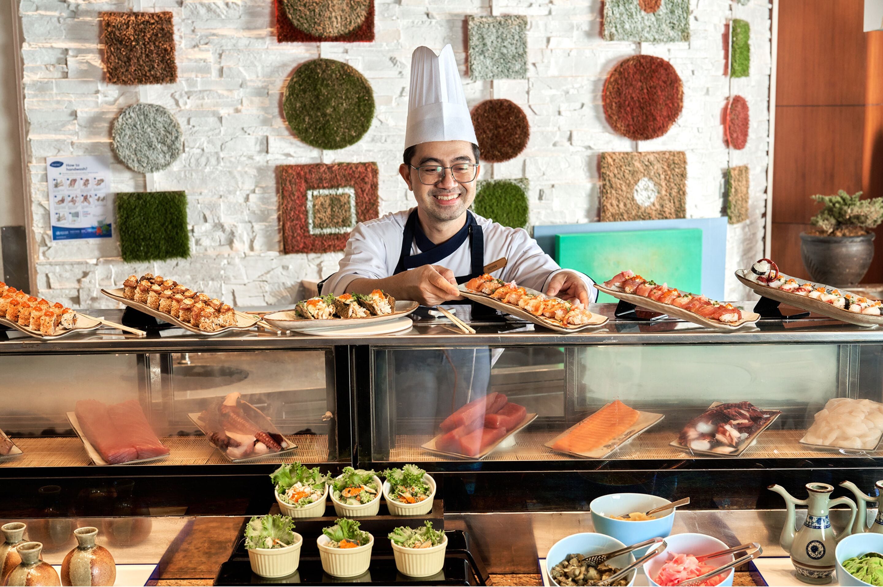 A chef in a white hat and apron behind a counter with various dishes and food items displayed.
