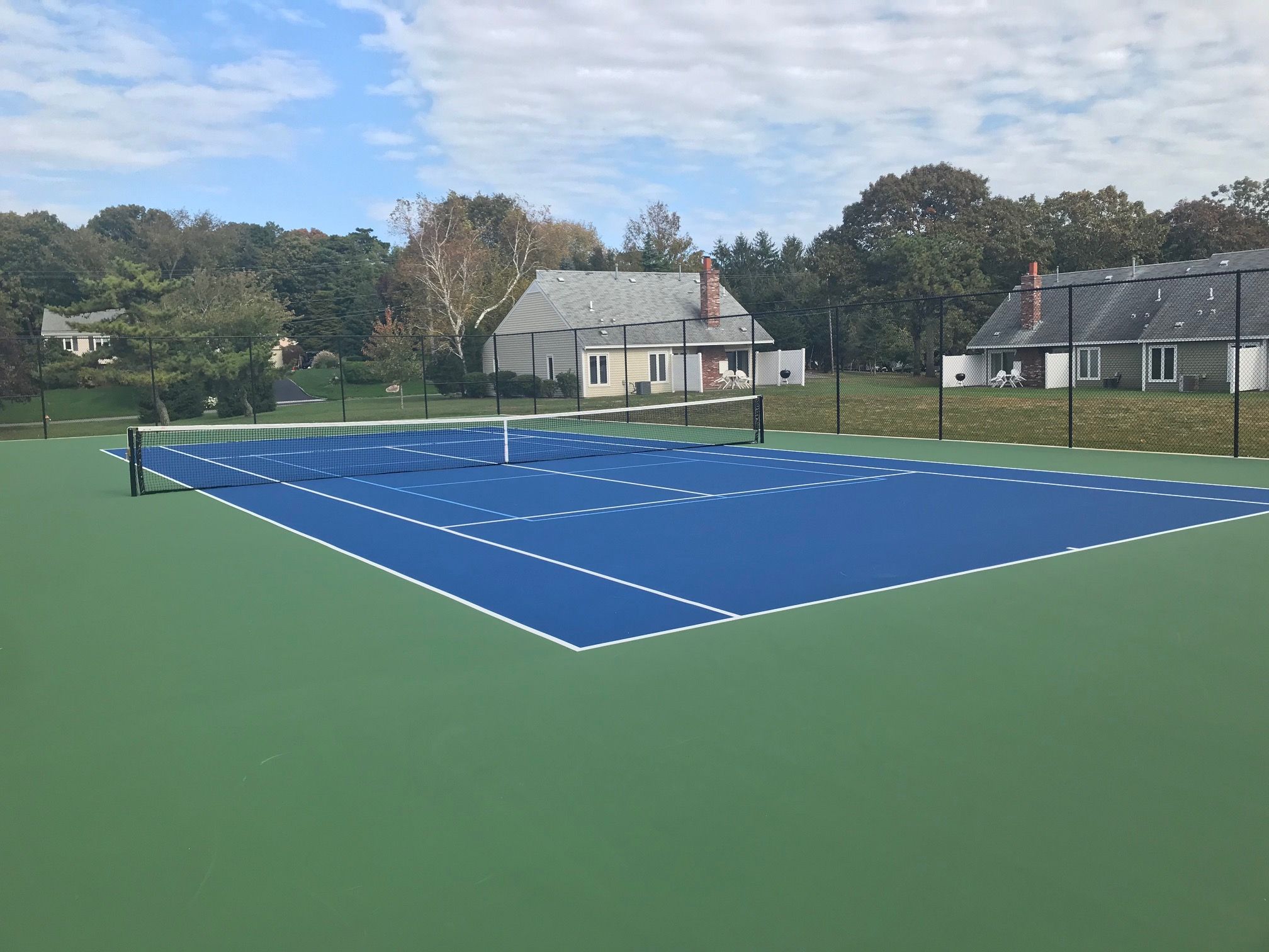 Una cancha de tenis al aire libre con red, rodeada por una valla, y casas al fondo.