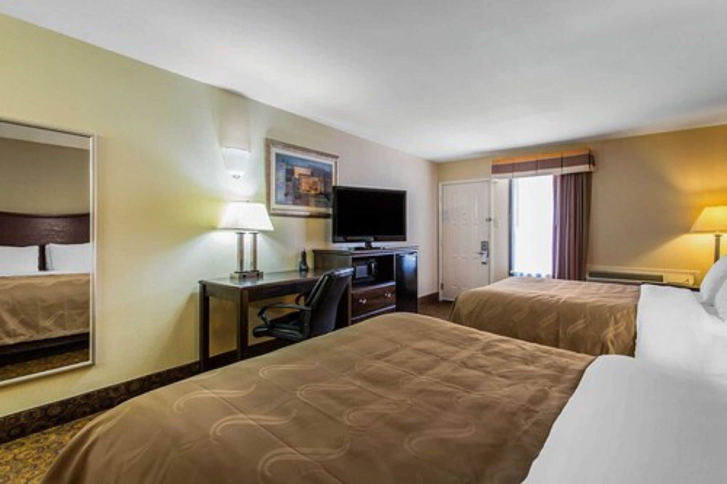 A hotel room with two beds, a desk with a chair, a television, and a window with curtains.