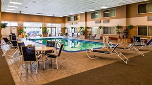 An indoor swimming pool with lounge chairs and tables, surrounded by windows and potted plants.