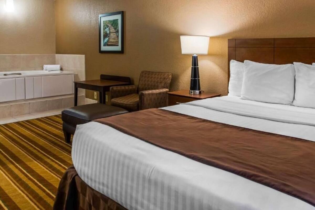 A hotel room with a large bed, a small table, a chair, a desk, a lamp, and a bathroom area.
