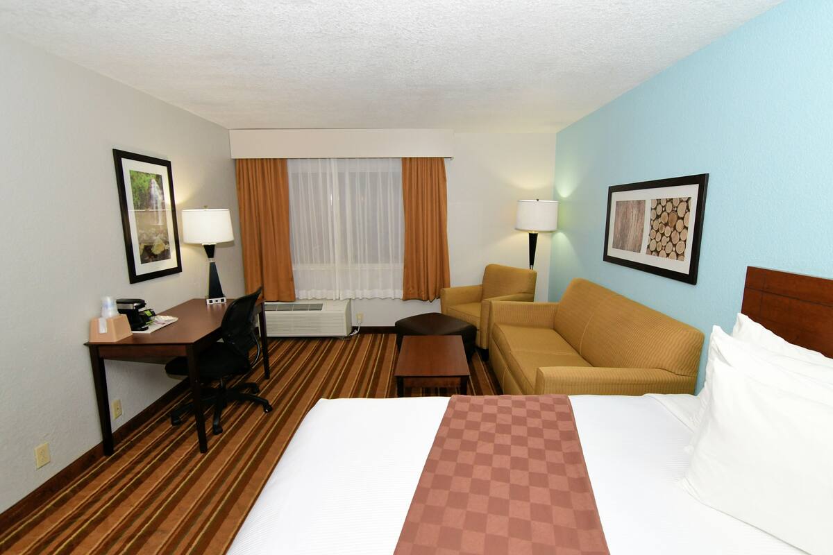 A hotel room with a bed, a desk with a chair, a sofa, a small table, a lamp, and a window with curtains.