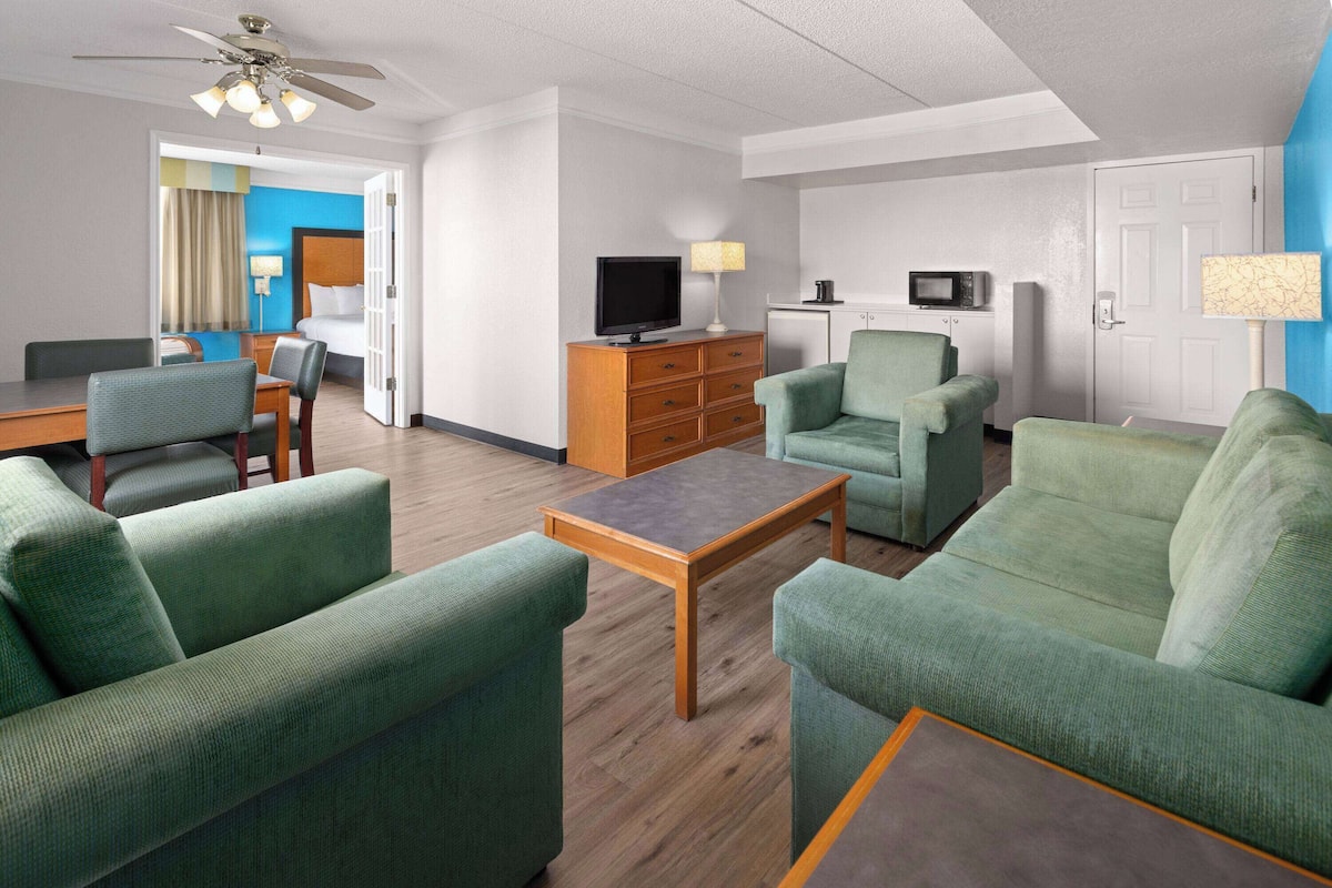 Une chambre d’hôtel dotée d’un coin salon comprenant des canapés recouverts d’un tissu vert, une table basse en bois, un téléviseur à écran plat, une lampe et un four à micro-ondes.