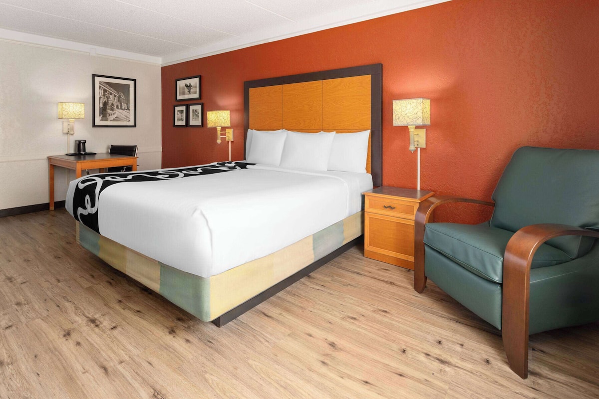 Une chambre d’hôtel avec un grand lit, un fauteuil vert, une table de chevet en bois et un bureau avec une lampe.