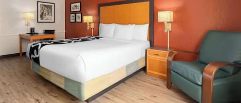 Quarto de hotel com uma cama grande, uma poltrona verde, uma mesa de cabeceira de madeira e uma escrivaninha com abajur.