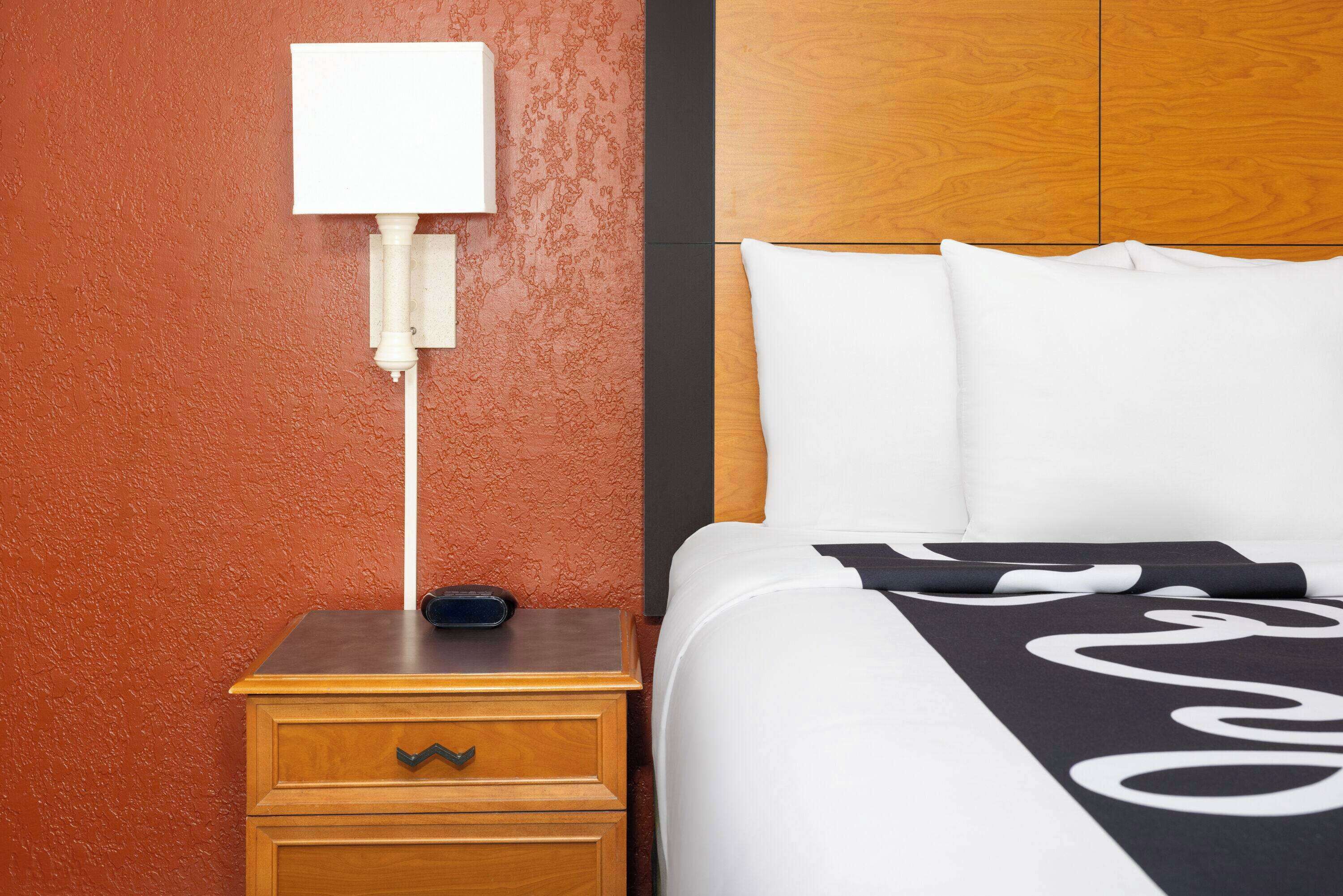 Une chambre d’hôtel avec une table de chevet en bois, une table de chevet blanche, un lit avec une literie blanche et un plaid noir et blanc, ainsi qu’une lampe fixée au mur.