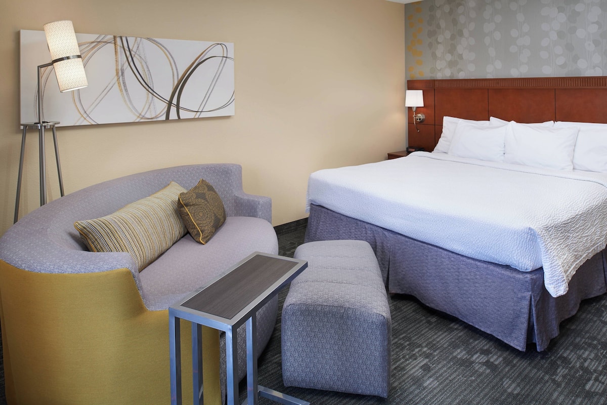 A hotel room with a bed, a sofa, an ottoman, a standing lamp, and a wall art piece.
