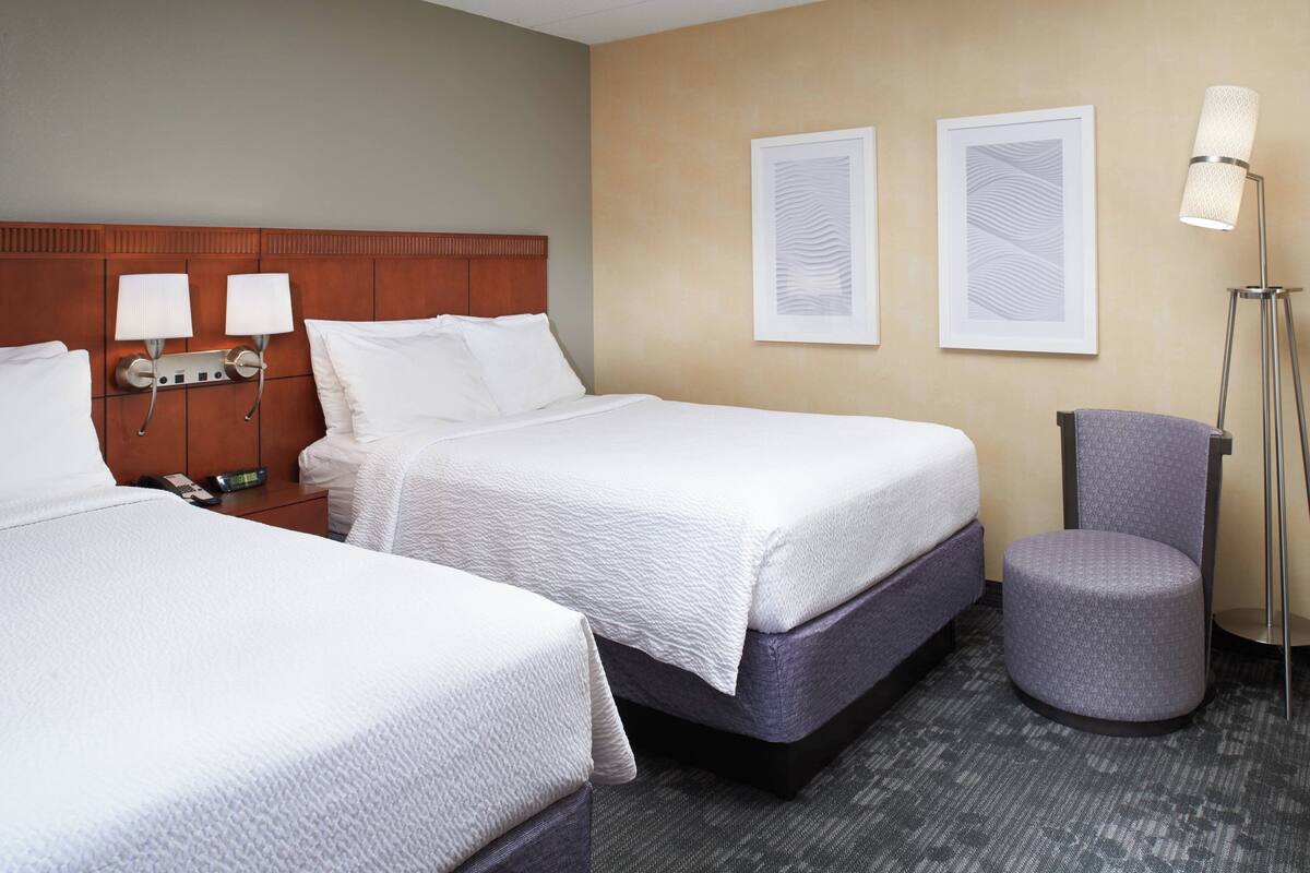A hotel room with two beds, a nightstand with a phone and lamp, and a chair.