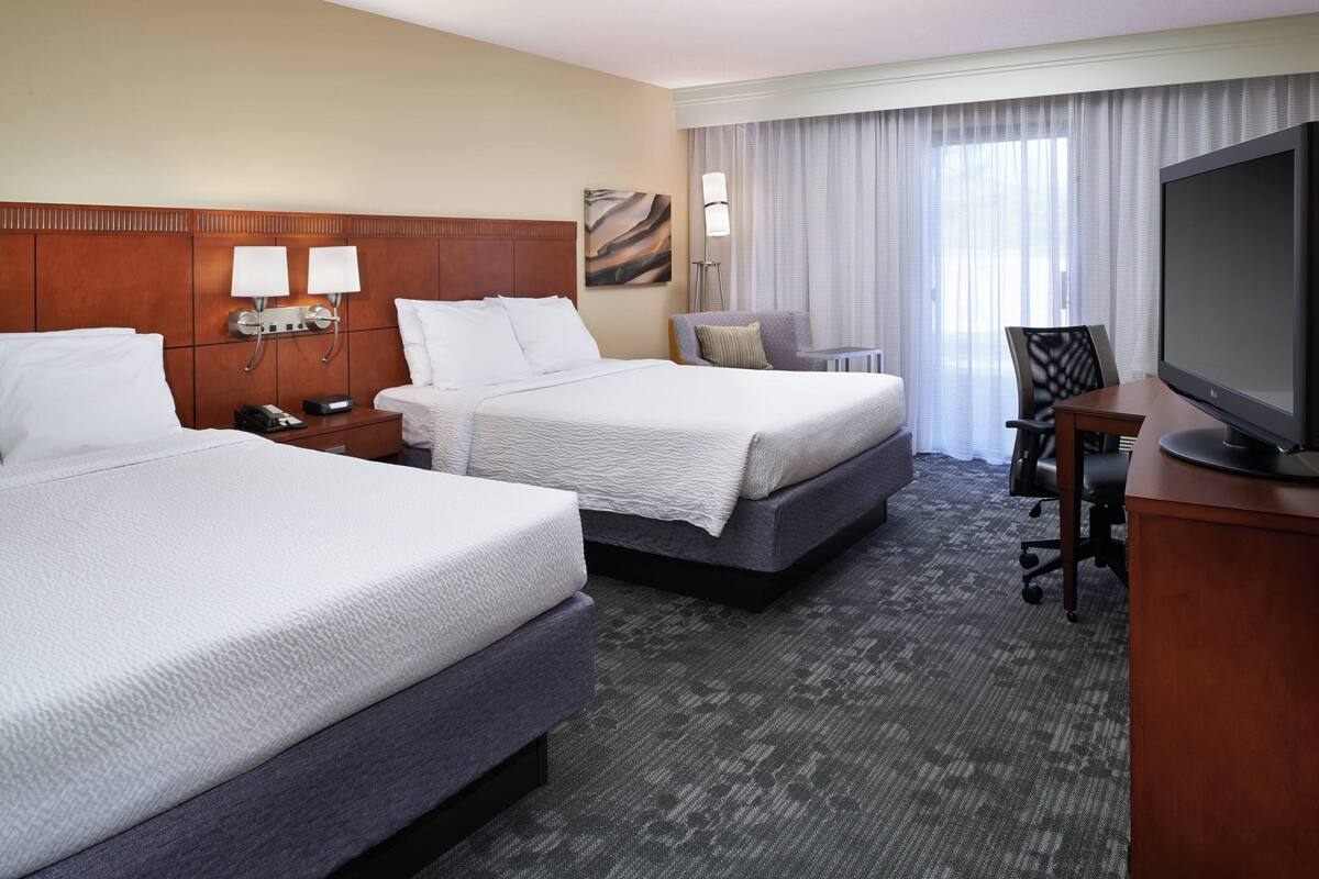 A hotel room with two beds, a desk, a television, and a window with curtains.