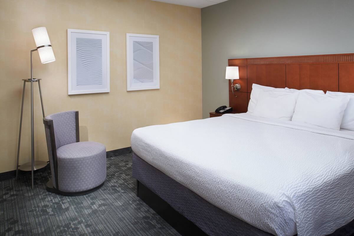 A hotel room with a large bed, a chair, two framed artworks, a floor lamp, and a bedside table with a lamp.