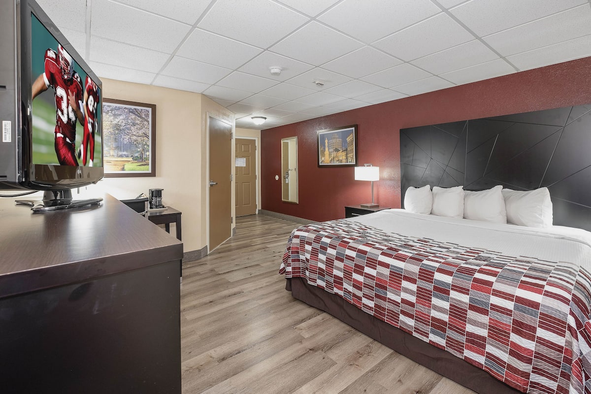 Una habitación de hotel con una cama grande, un escritorio con televisor, un cuadro en la pared y una puerta que da a otra habitación.