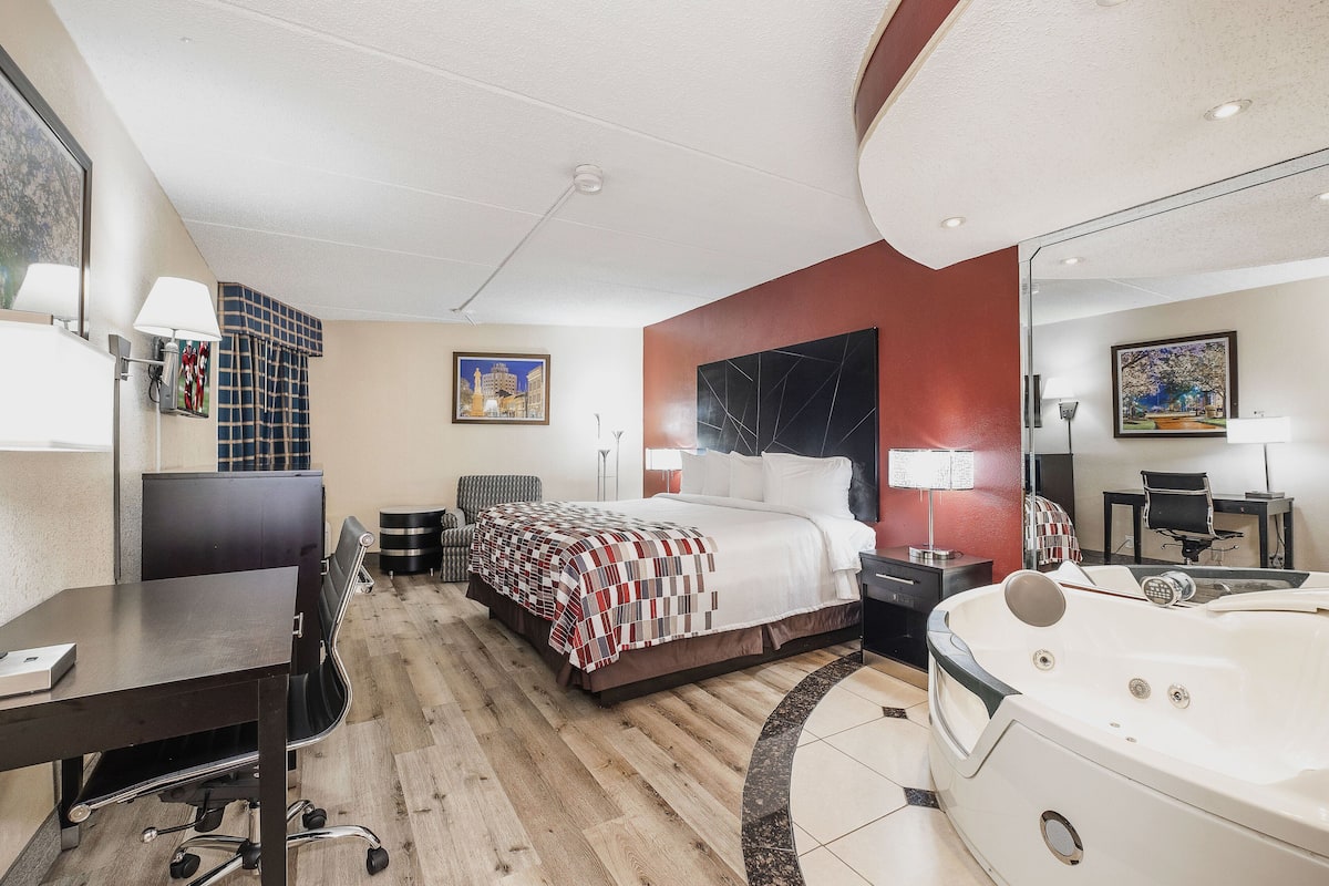 Habitación de hotel con cama, escritorio, silla, lámpara de escritorio, mesita de noche, espejo, ventana grande y bañera.