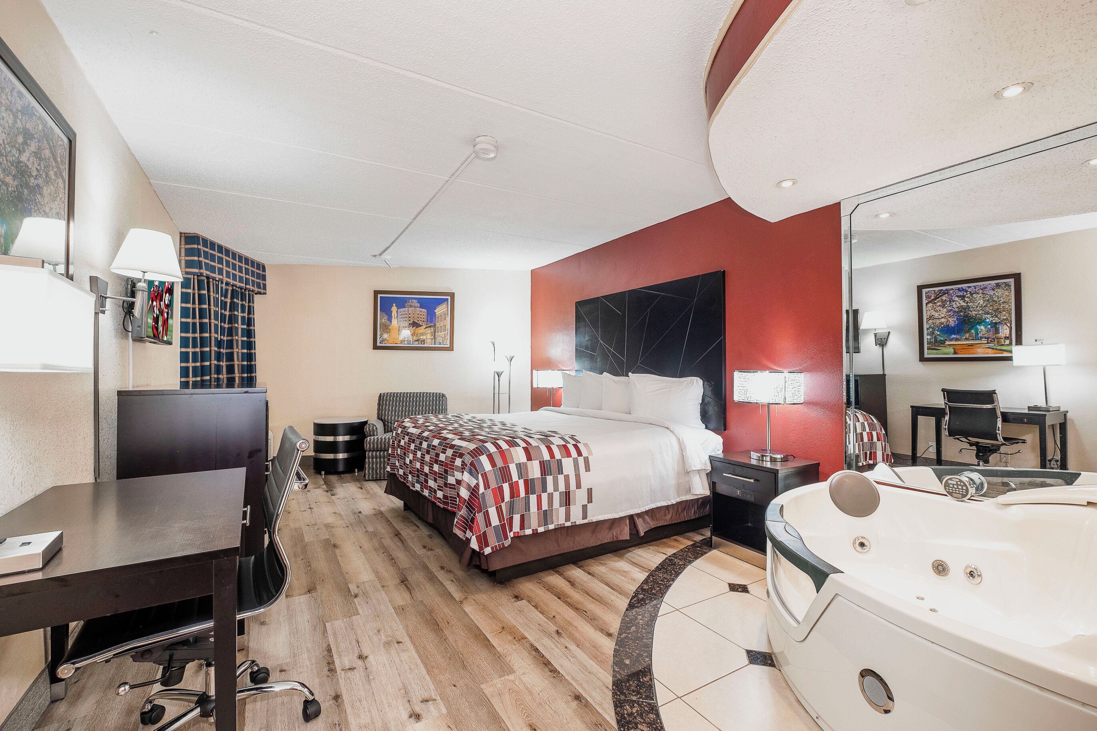 Une chambre d’hôtel comprenant un lit, un bureau, une chaise, une lampe de bureau, une table de chevet, un miroir, un tableau, une fenêtre et une baignoire.