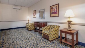 A hotel room with patterned carpet, a wooden side table with a lamp, a wooden chair, a small wooden cabinet, and a wall-mounted mirror. - Red Roof Inn & Suites Macon (Macon)