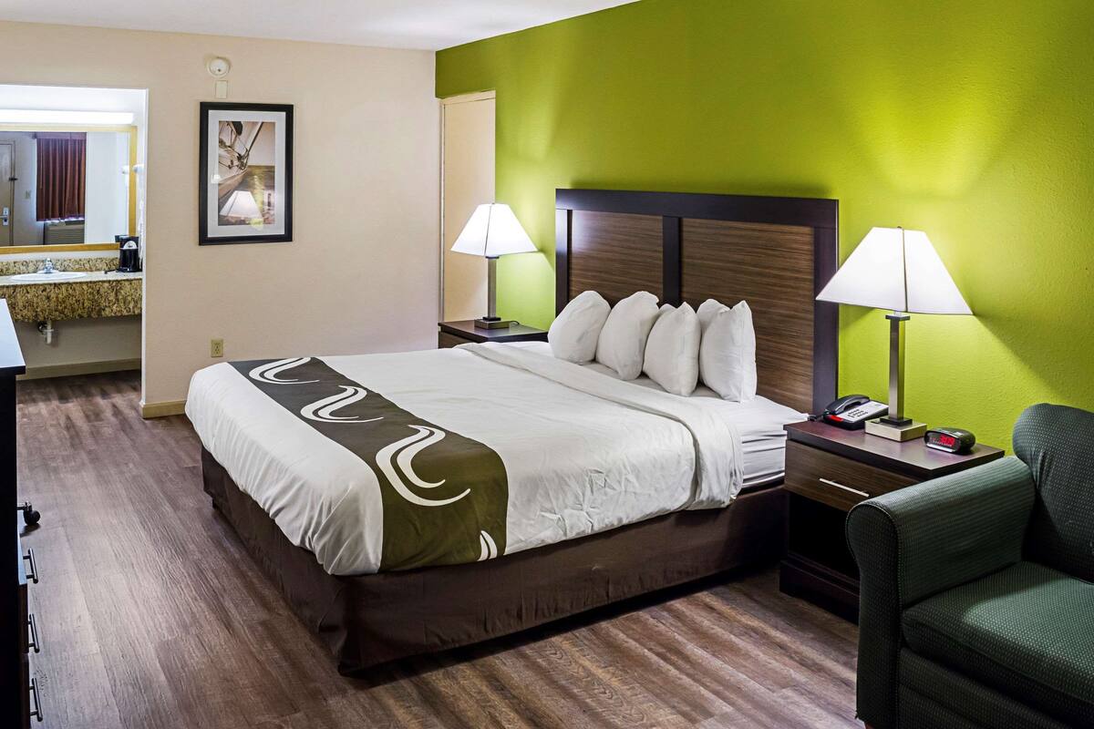 A hotel room with a large bed, two bedside lamps, a nightstand with a phone and alarm clock, a green accent wall, and a bathroom visible in the background.