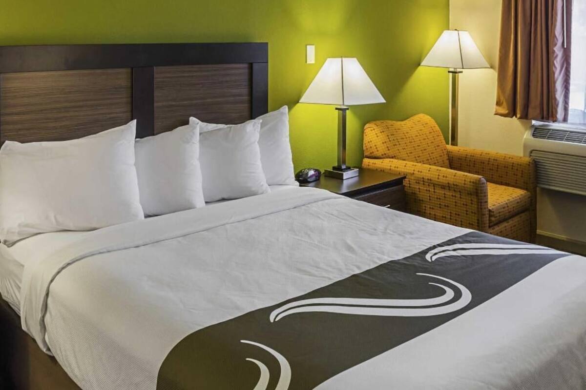 A hotel room with a bed, two lamps, a chair, and a window with curtains.