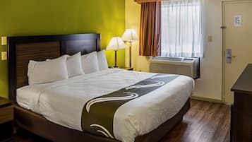 A neatly made bed with white linens and a decorative pillow, a bedside lamp, and a window with curtains.