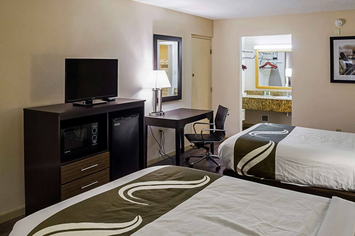 A hotel room with two beds, a desk with a television, a desk lamp, a chair, and a mirror.