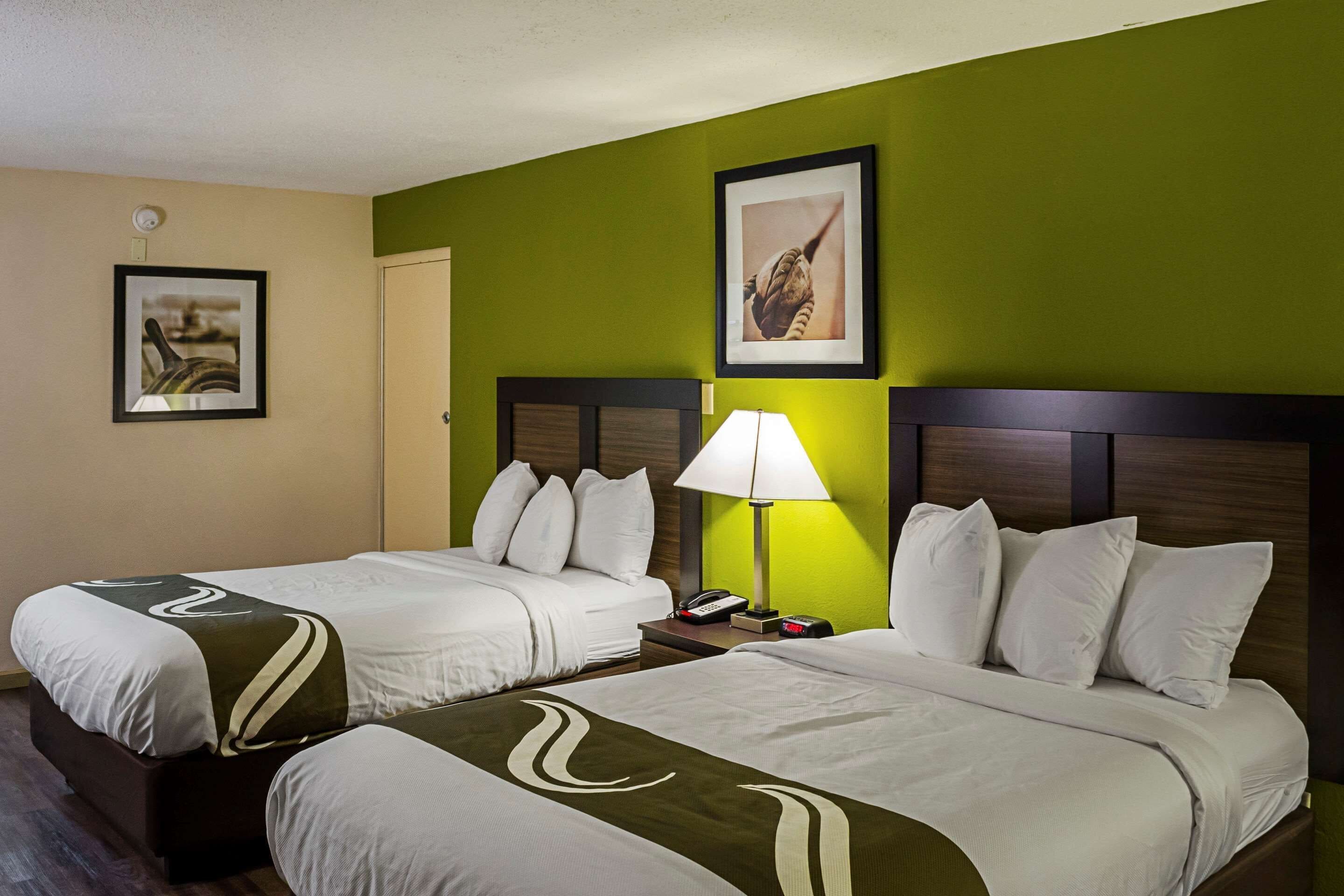 A hotel room with two beds, a nightstand with a lamp, and artwork on the walls.