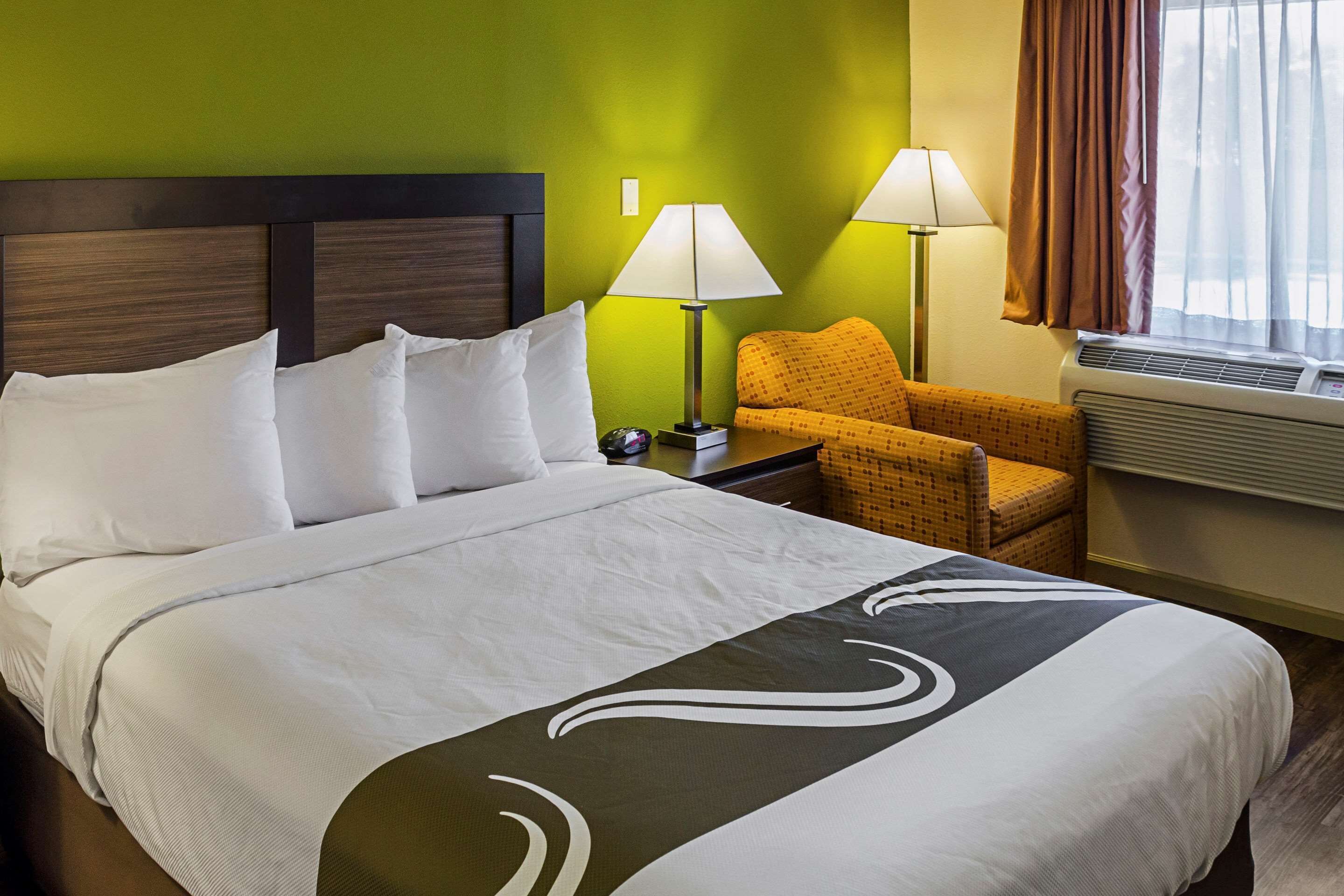 Una habitación de hotel con una cama grande, dos lámparas de noche, una silla y un ventanal con cortinas.