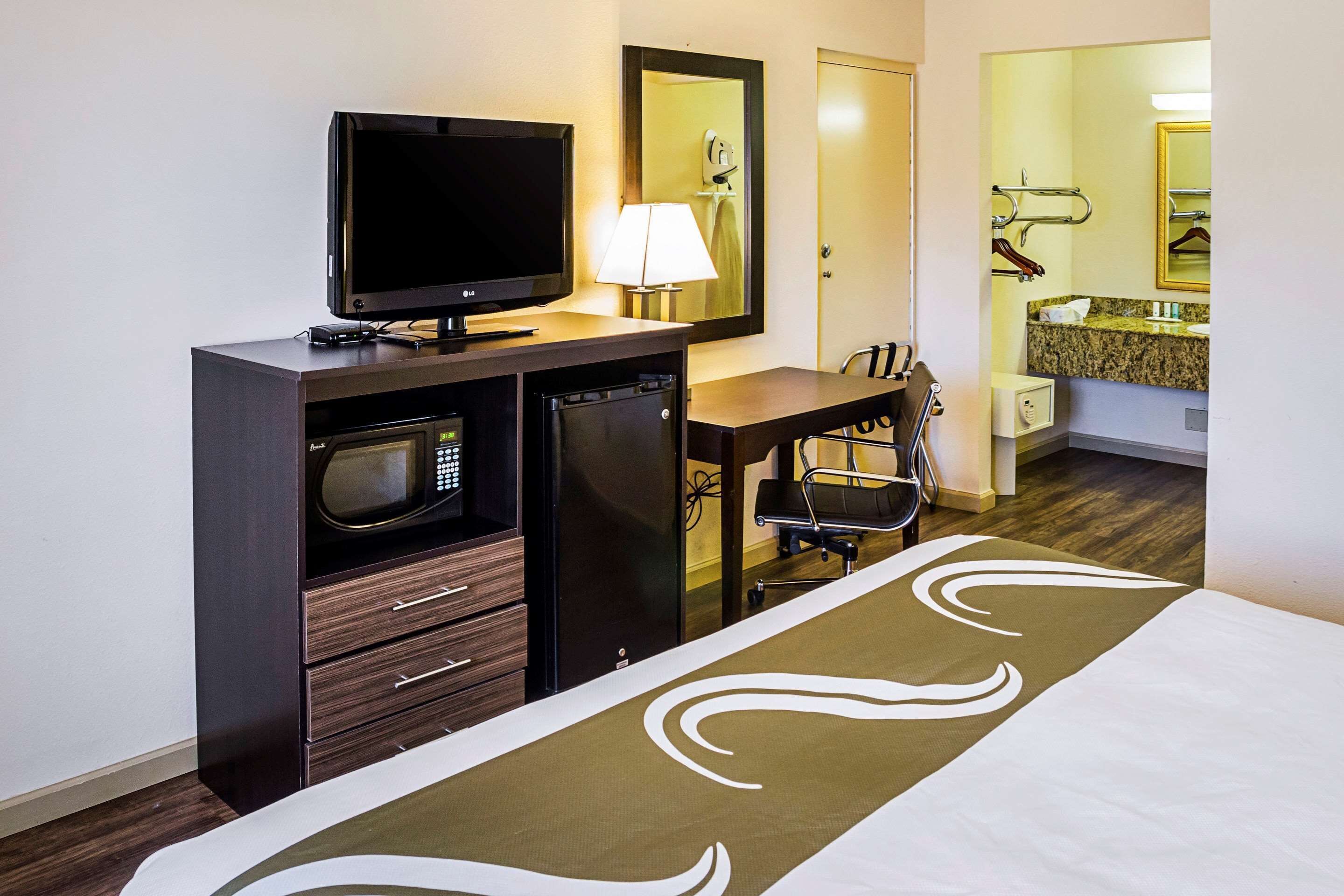 Ein Hotelzimmer mit Bett, Fernseher, Mikrowelle, Kühlschrank, Schreibtisch und einem Badezimmer mit Waschbecken und Spiegel.