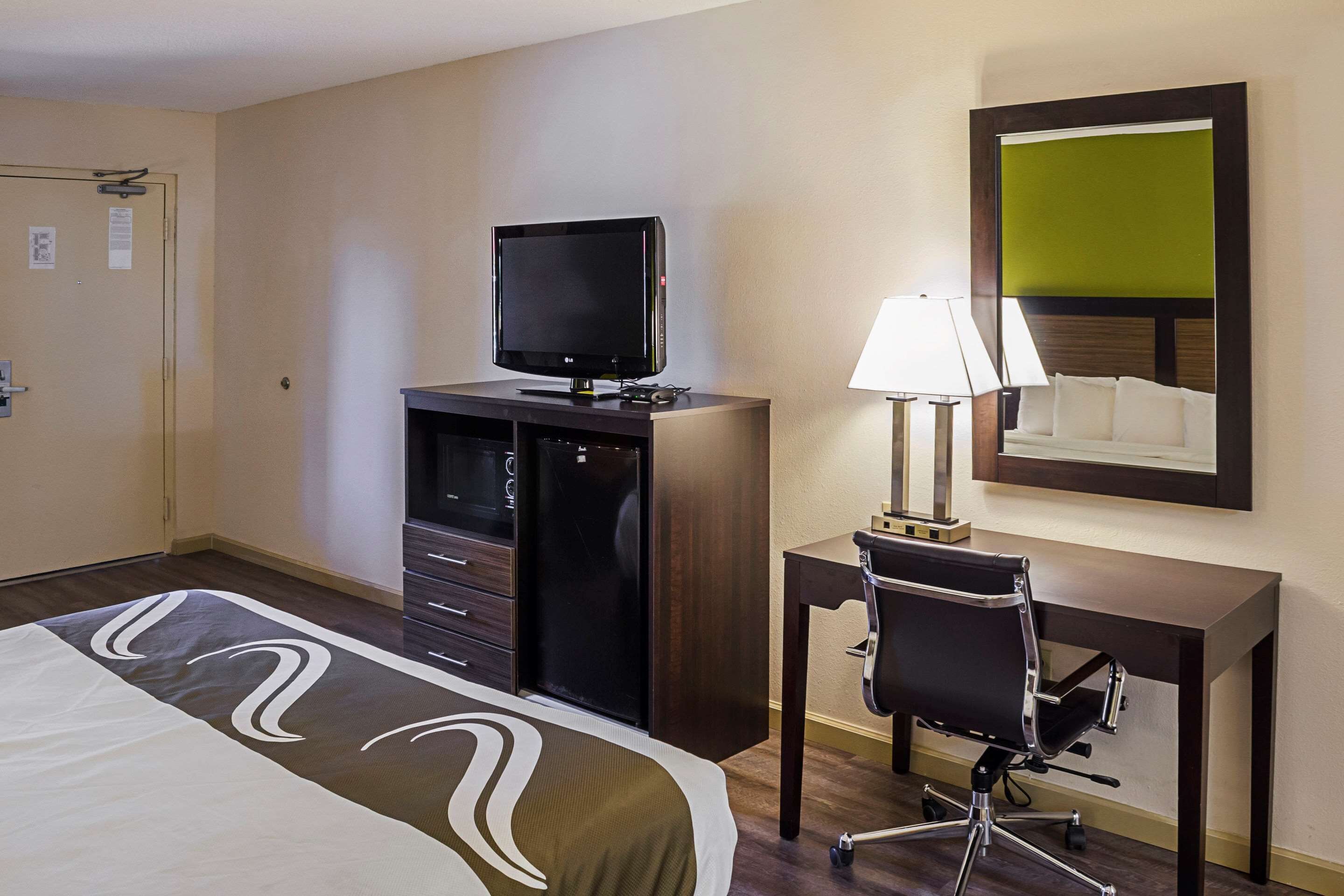 Een hotelkamer met een bed, een bureau met een computer en lamp, een televisie op een dressoir, en een spiegel aan de muur.