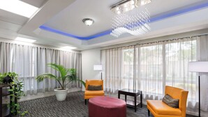 Habitación de hotel con diseño moderno, que incluye un ventanal con cortinas translúcidas, una planta en maceta, un reposapiés rojo y dos sillones naranjas.