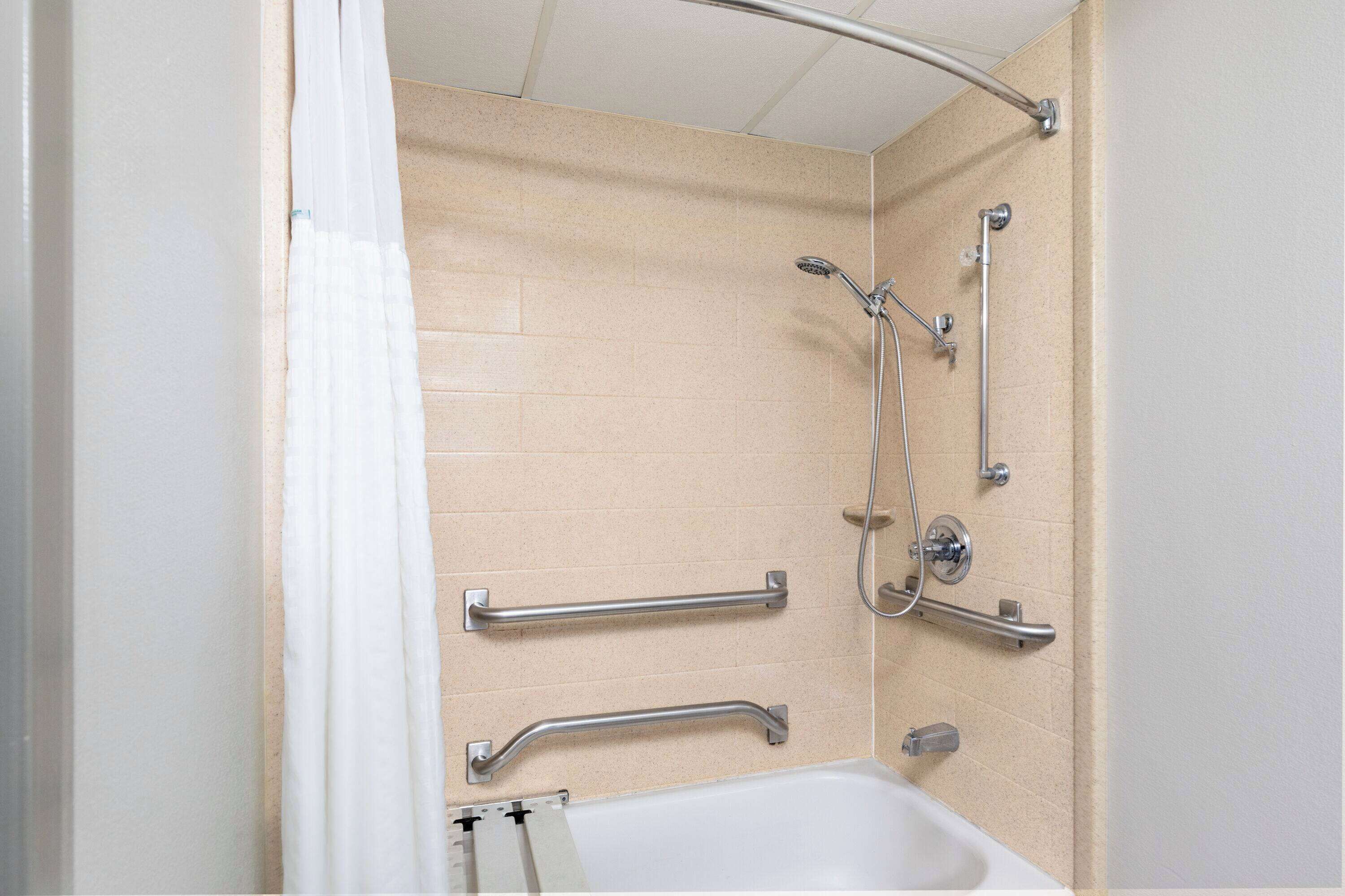 Un baño con baldosas color beige, una bañera blanca y una ducha con cabezal de ducha manual y barras de apoyo.
