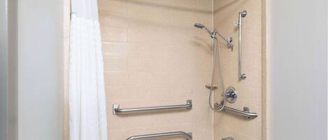 Un baño con baldosas color beige, una bañera blanca y una ducha con cabezal de ducha manual y barras de apoyo.