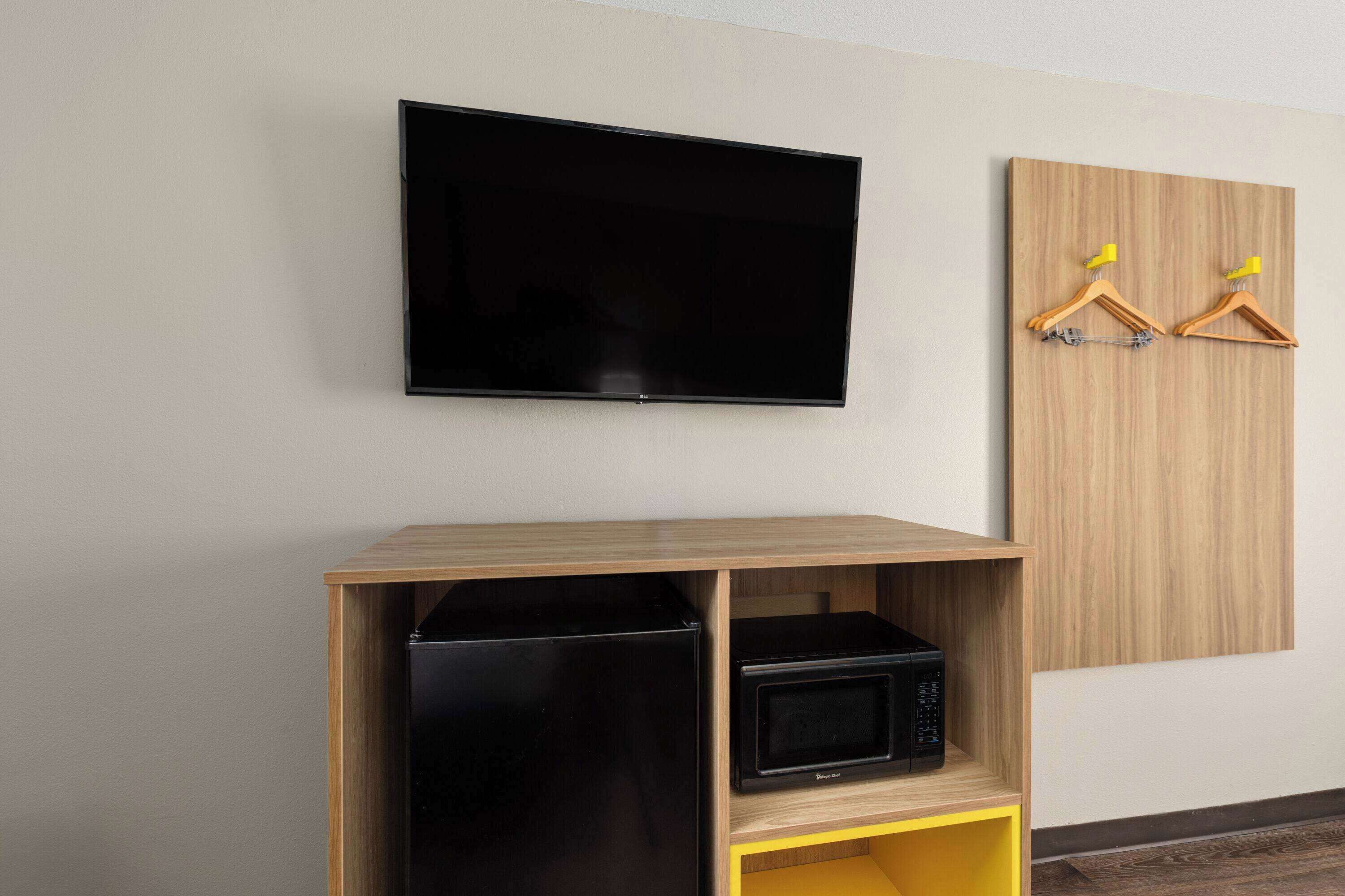Una habitación con una televisión de pantalla plana montada en la pared, un mueble de madera con un microondas y dos perchas de madera sobre un panel de madera.