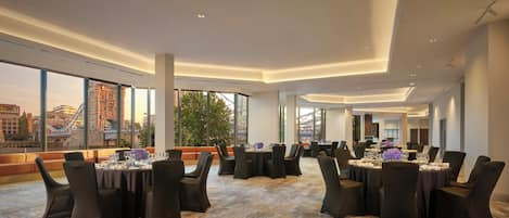 A spacious conference room with multiple tables and chairs, large windows offering a city view, and a modern ceiling design.