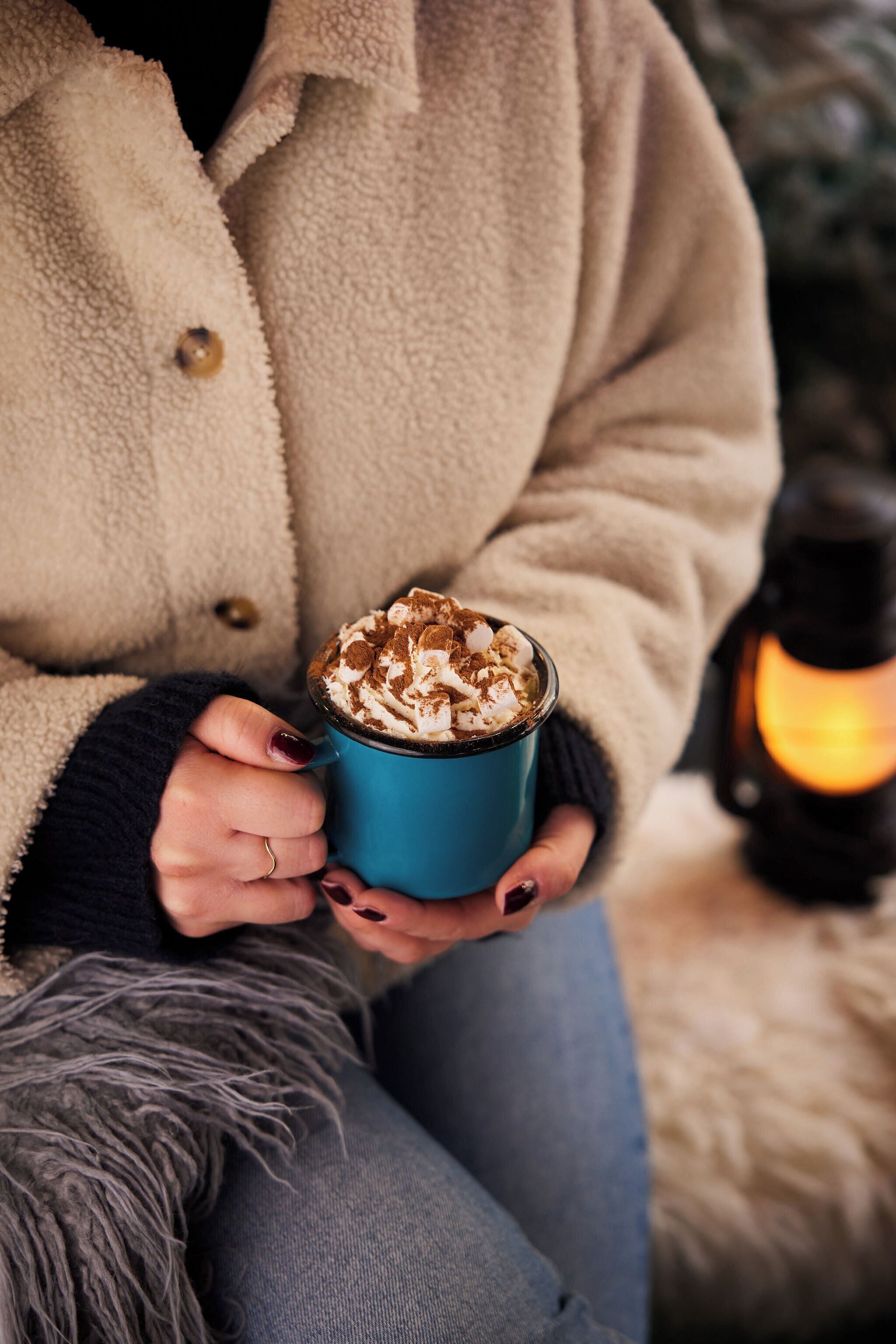 A person holding a mug with whipped cream, wearing a beige coat, and sitting near a lit lantern.