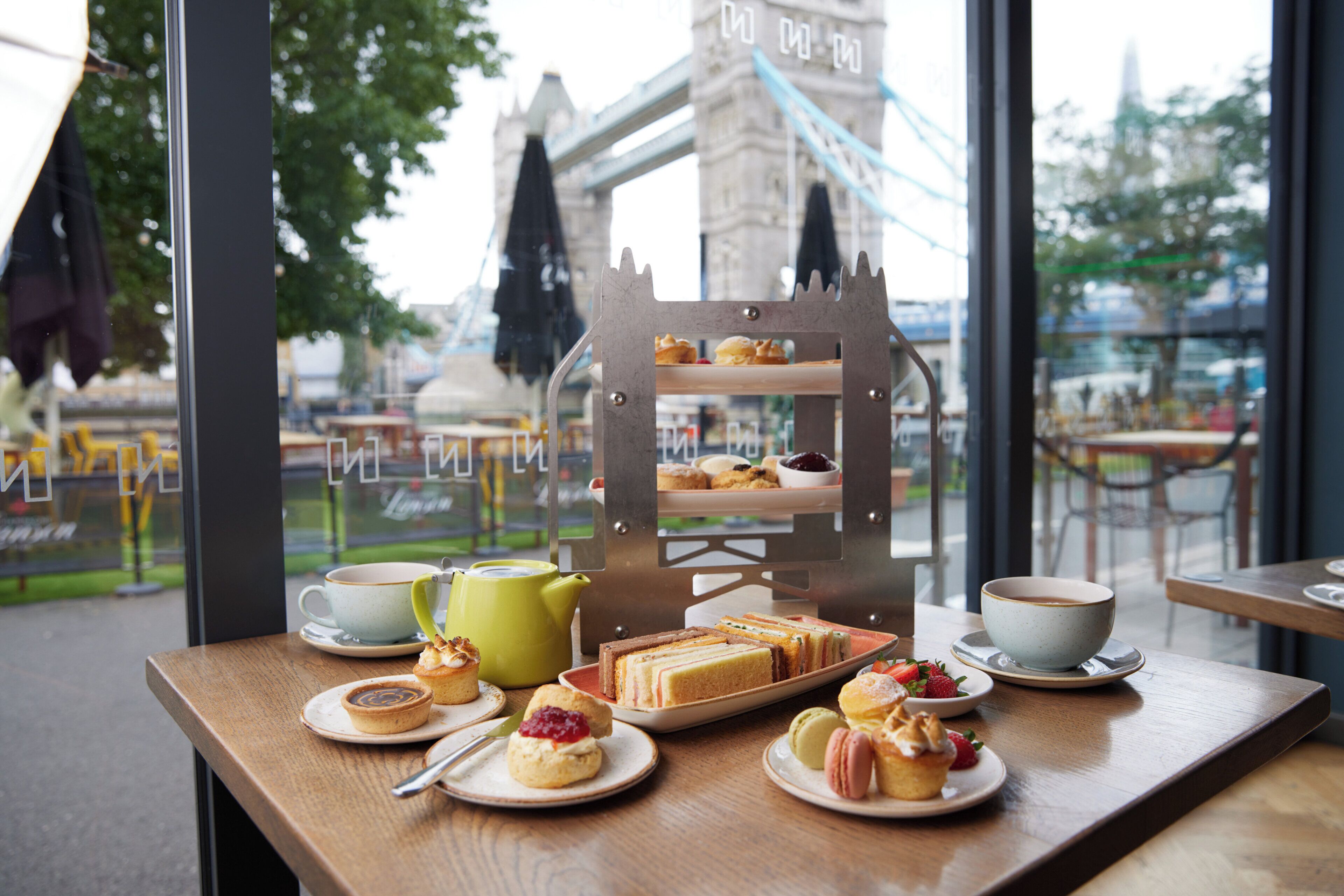 A table with pastries and a tiered stand of sandwiches, with a view of a bridge and greenery outside.