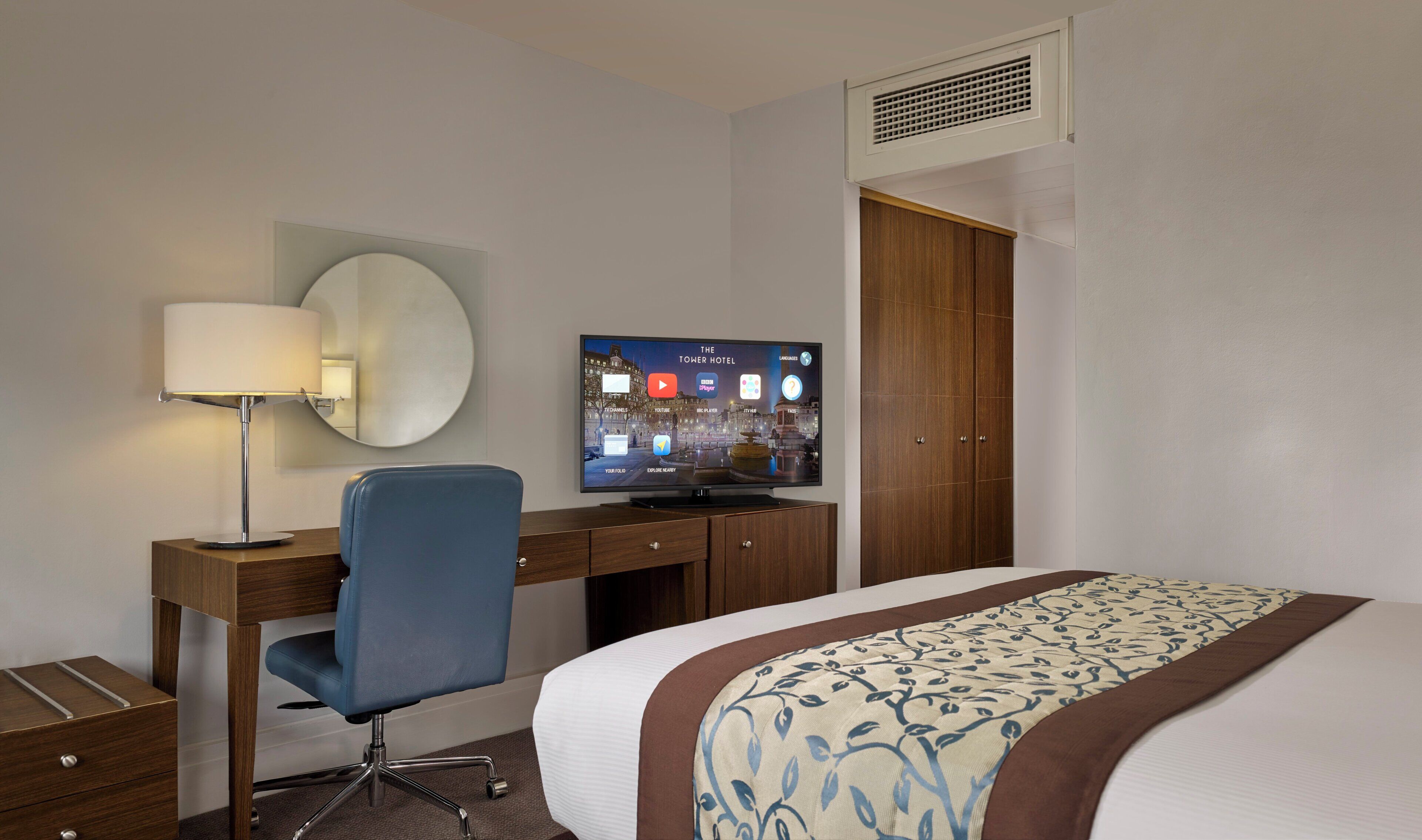 Et hotelværelse med en seng, et skrivebord med stol, et fjernsyn der viser en hotelapp og et klædeskab.