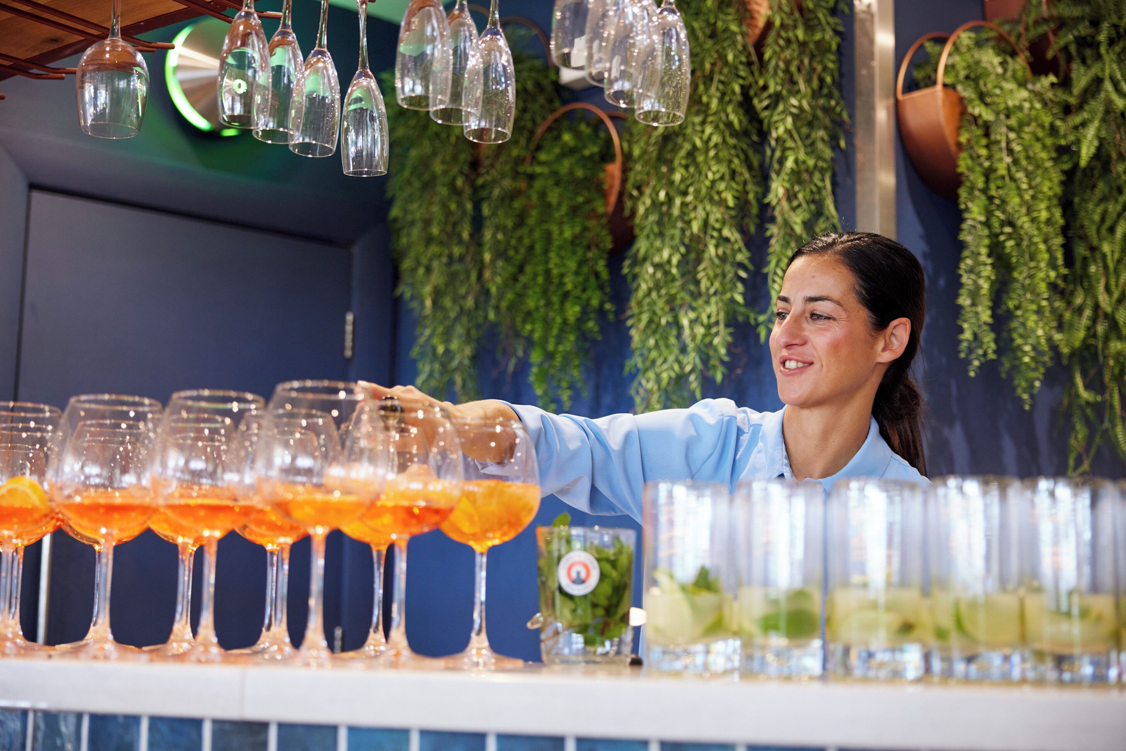 A bar area with multiple glasses of orange beverages, a bartender, and hanging light fixtures.