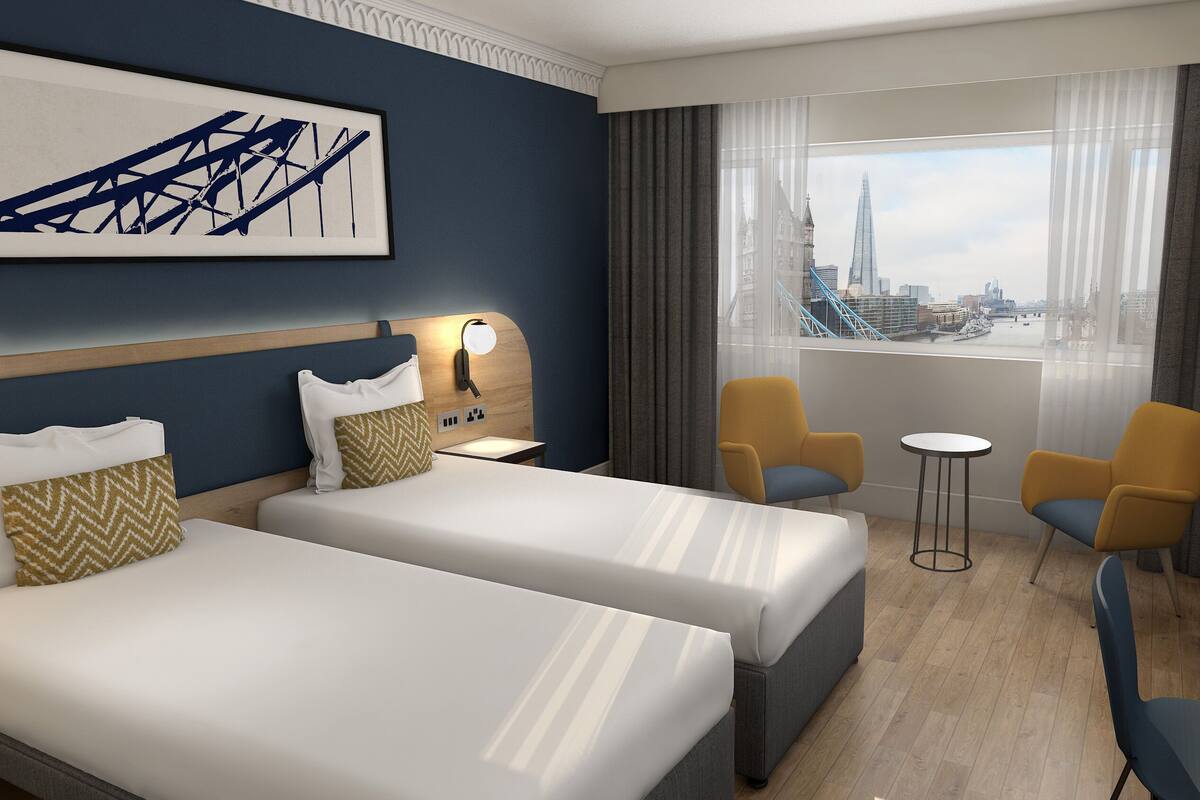 A hotel room with two beds, a desk, two chairs, a view of a cityscape, and a framed artwork on the wall.