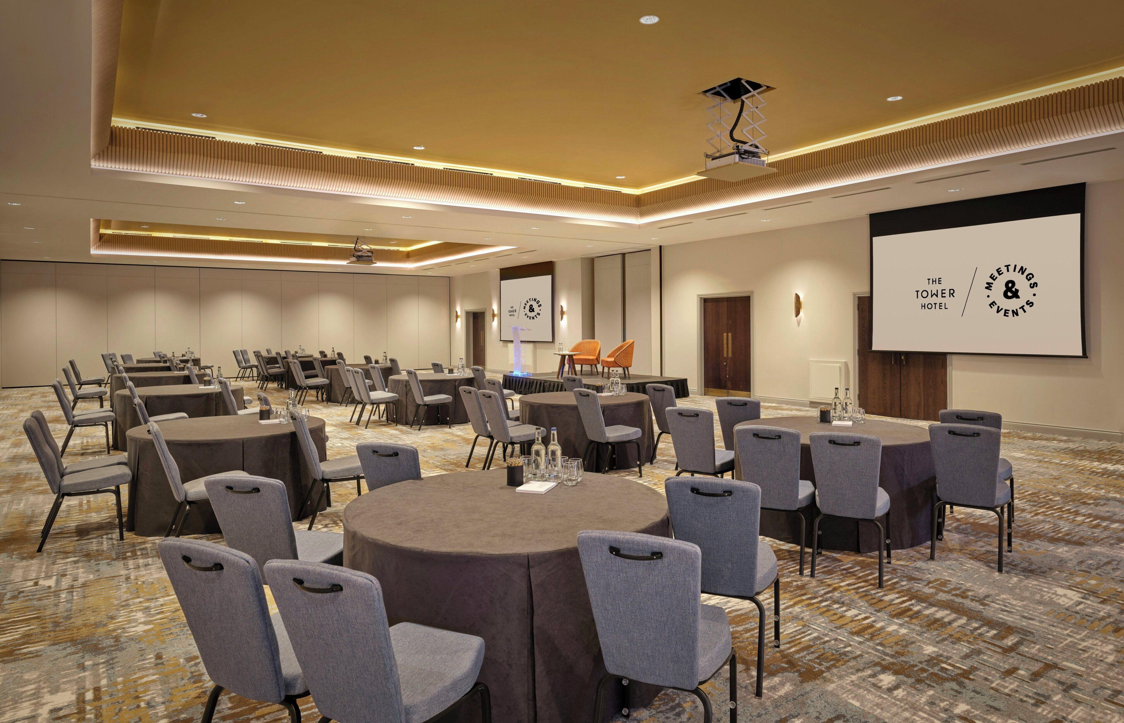 A conference room with multiple tables and chairs, a projector screen, and a carpeted floor.