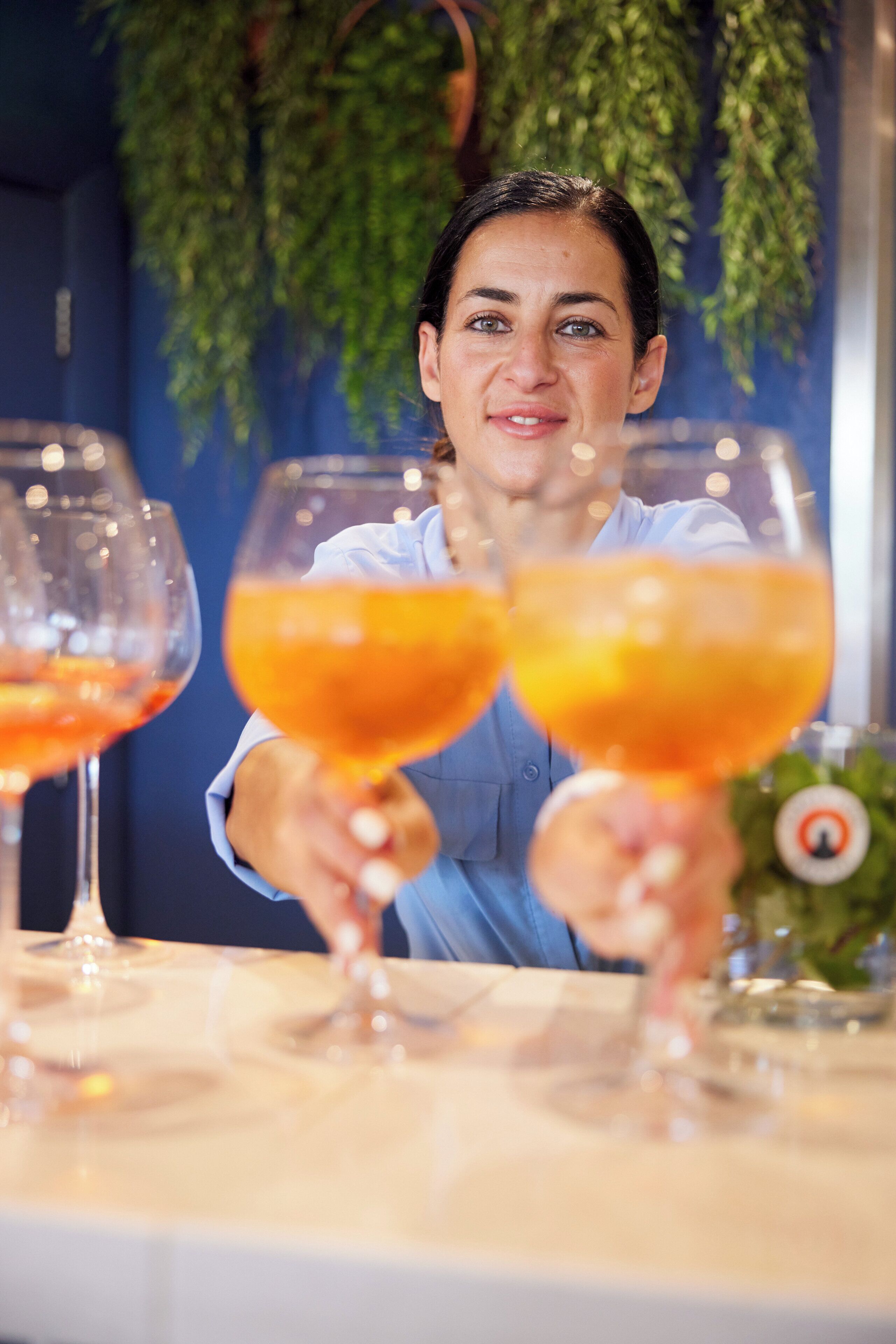 A person holding two wine glasses at a table with a blue wall and hanging greenery in the background.
