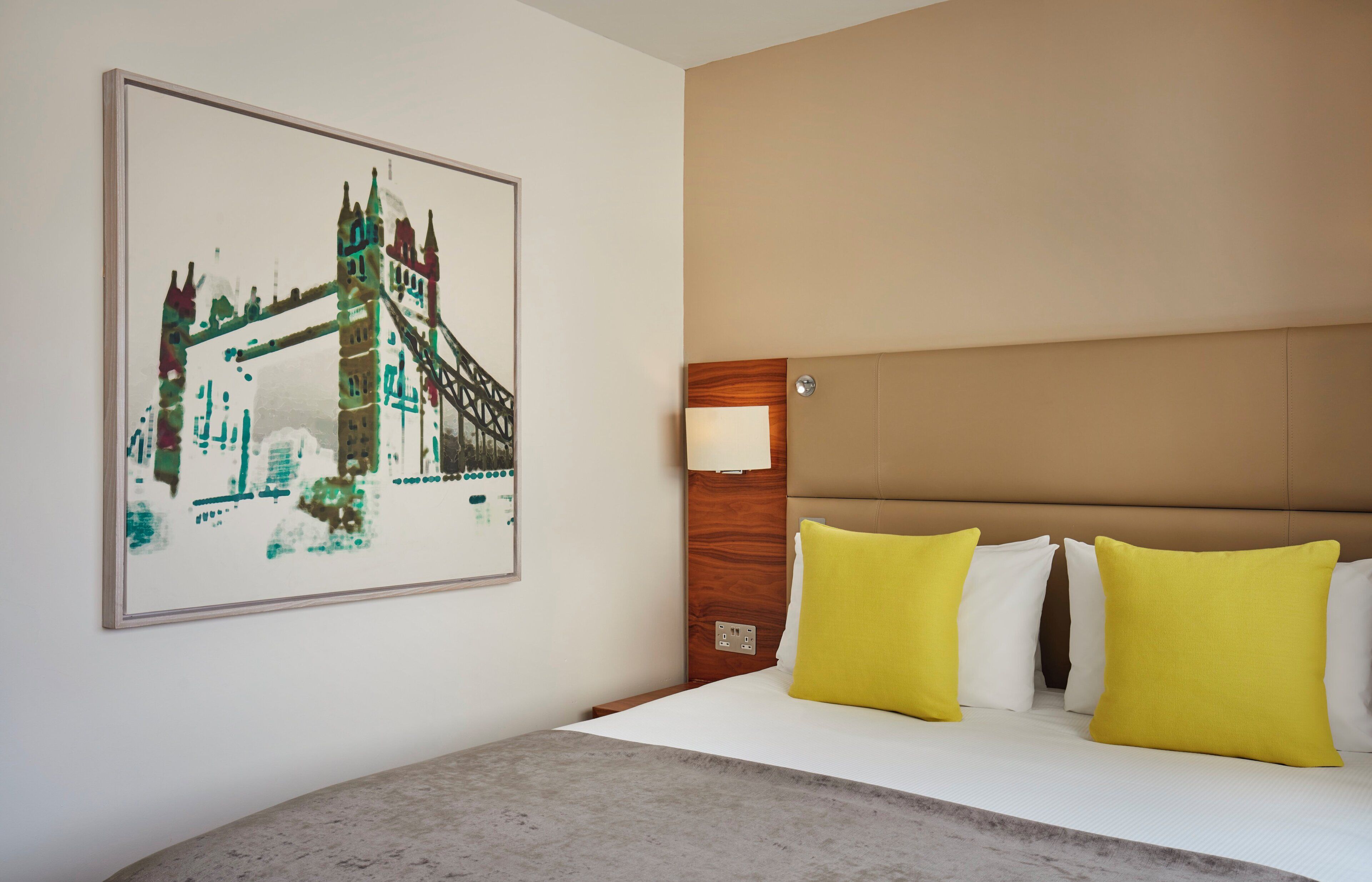 A hotel room with a bed, a headboard, a wall-mounted lamp, and a framed picture of a bridge.