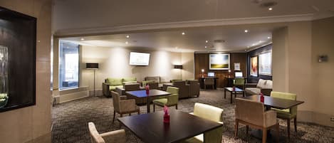 A hotel lobby with multiple seating areas, including sofas and tables, a TV screen, and a patterned carpet.